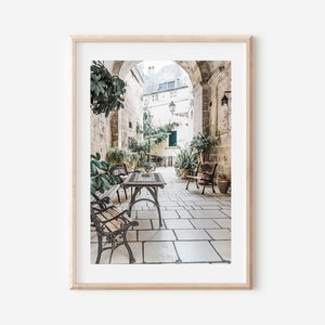 May include: A framed print of an outdoor cafe scene. The image features a stone-paved courtyard with tables, benches, and lush greenery. The scene is framed by a light-colored wooden frame.