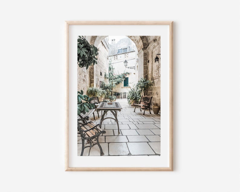 May include: A framed print of an outdoor cafe scene. The image features a stone-paved courtyard with tables, benches, and lush greenery. The scene is framed by a light-colored wooden frame.