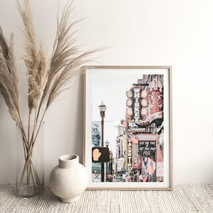 May include: A framed print of a city street scene with neon signs, including a sign that says "Whiskey Saloon" and a boot-shaped sign. The signs are lit up and there are other buildings and people in the background.
