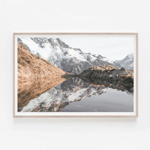 Puede incluir: Una fotograf&iacute;a panor&aacute;mica de una cordillera reflejada en un lago tranquilo. Las monta&ntilde;as est&aacute;n cubiertas de nieve y el lago es de un azul profundo. La imagen est&aacute; enmarcada en un marco de madera claro.