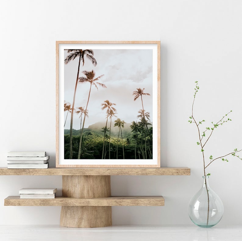 May include: A framed print of a tropical landscape with palm trees and a lush green hillside. The print is in a light wood frame and is hanging on a white wall.