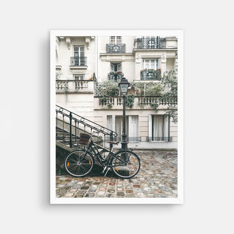 May include: A black bicycle is parked on a cobblestone street in front of a white building with a black wrought iron balcony. The building has several windows and a street lamp is next to the bicycle.