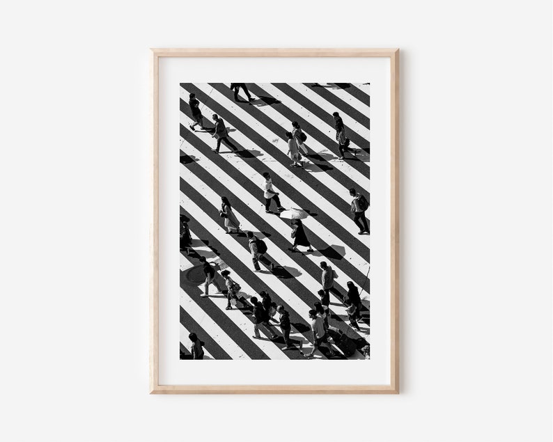 May include: Black and white photo of people walking across a crosswalk with diagonal stripes. The photo is taken from above, and the people's shadows are visible on the ground.