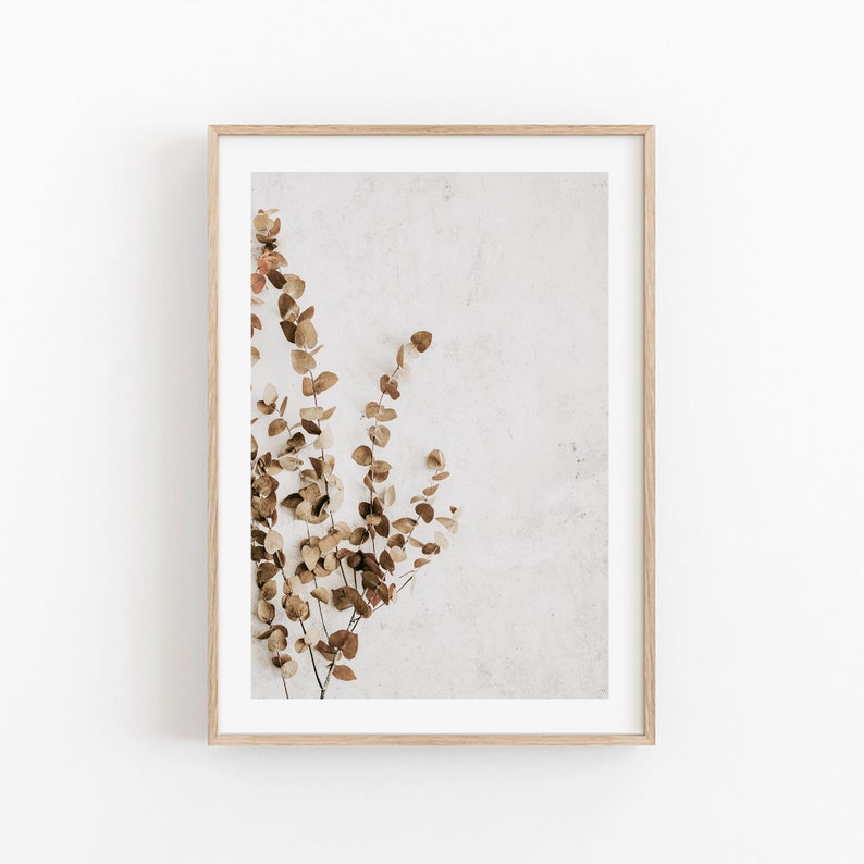 May include: A framed print showing a sprig of dried eucalyptus leaves in brown and tan hues against a textured, off-white backdrop. The print is set within a light-coloured wooden frame, offering a minimalist design.