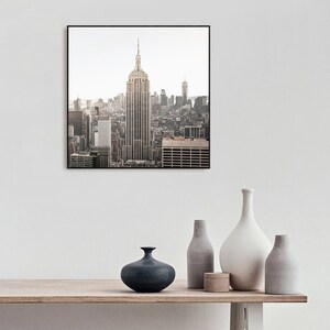 May include: A framed photograph of the New York City skyline with the Empire State Building in the center. The photo is in a black frame and is hanging on a white wall above a wooden table with vases.