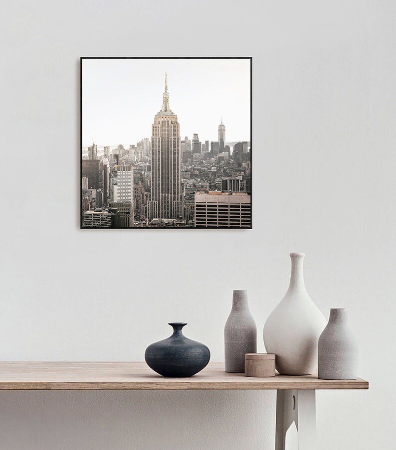 May include: A framed photograph of the New York City skyline with the Empire State Building in the center. The photo is in a black frame and is hanging on a white wall above a wooden table with vases.