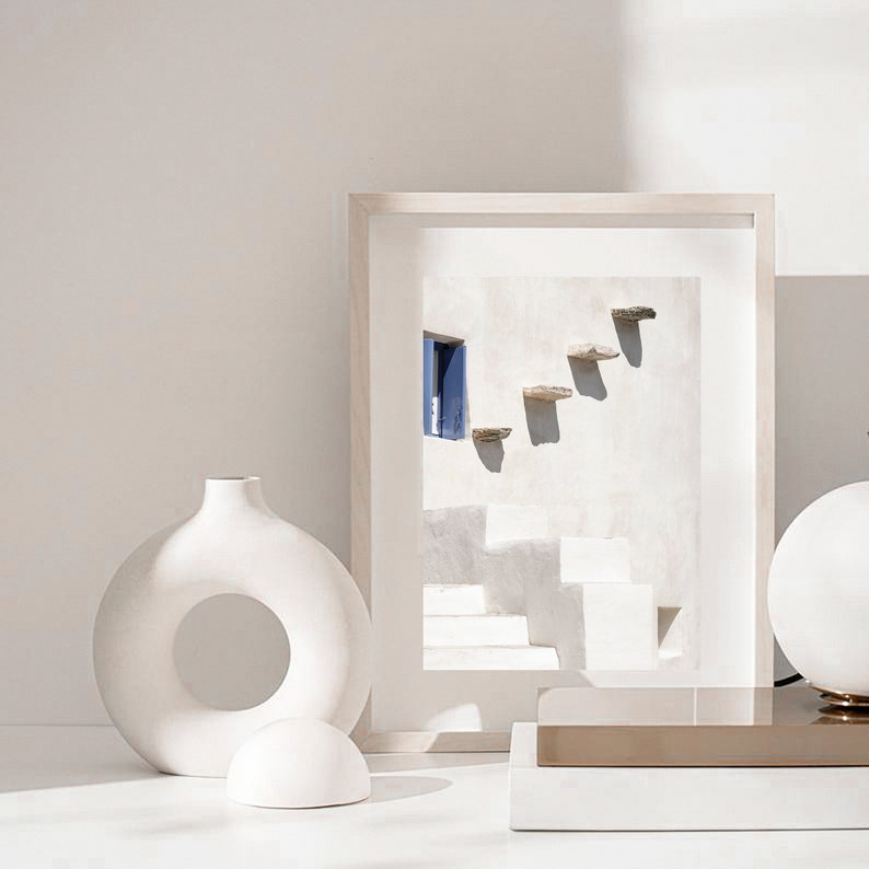 May include: White ceramic vase with a circular opening, a white ceramic sphere, and a framed photograph of a white building with a blue door and steps. The photograph is on a white surface with a gold metal base.