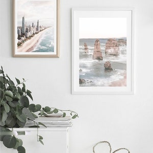 May include: Two framed photographs of coastal scenes. The first photo shows a city skyline with buildings along a sandy beach. The second photo shows a rocky coastline with large rock formations in the ocean. The photos are hanging on a white wall above a white cabinet with a stack of magazines on top.