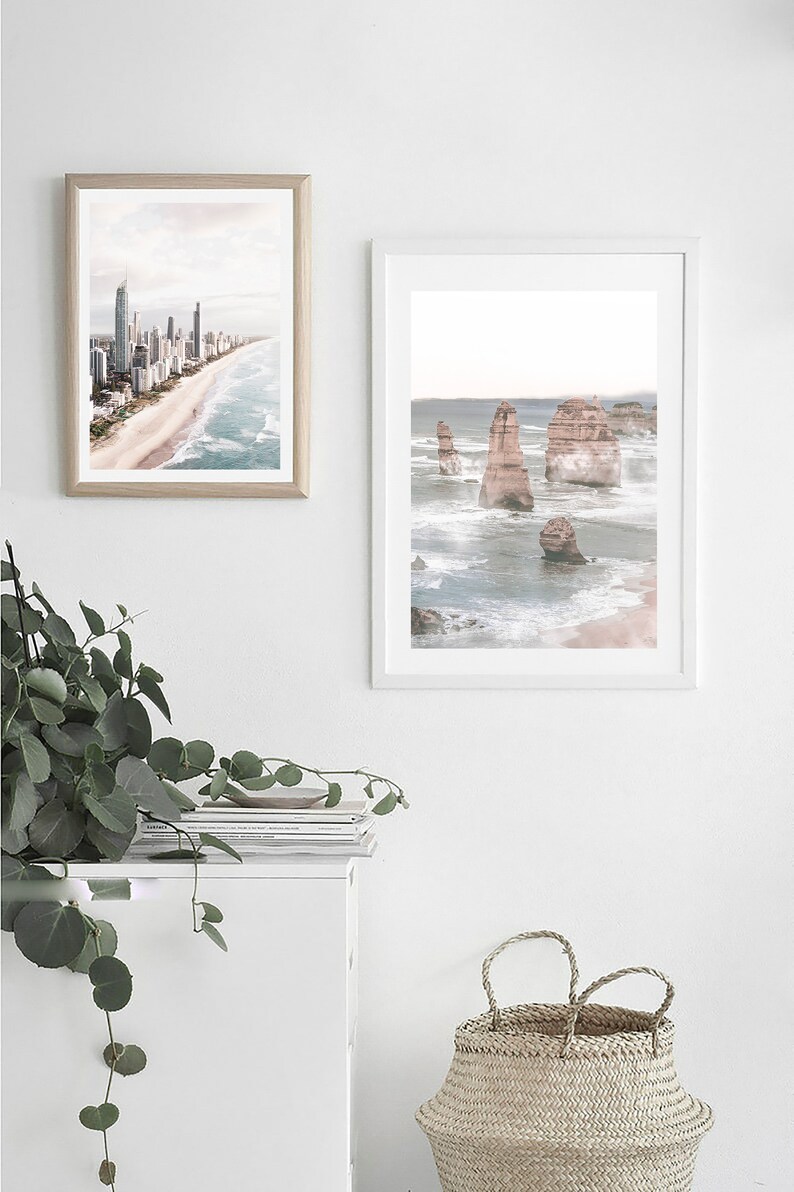 May include: Two framed photographs of coastal scenes. The first photo shows a city skyline with buildings along a sandy beach. The second photo shows a rocky coastline with large rock formations in the ocean. The photos are hanging on a white wall above a white cabinet with a stack of magazines on top.