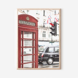 May include: A red telephone booth with the word "TELEPHONE" on the front, located on a street in London, England. A black taxi cab is driving past the booth, and there is a traffic light in the background.