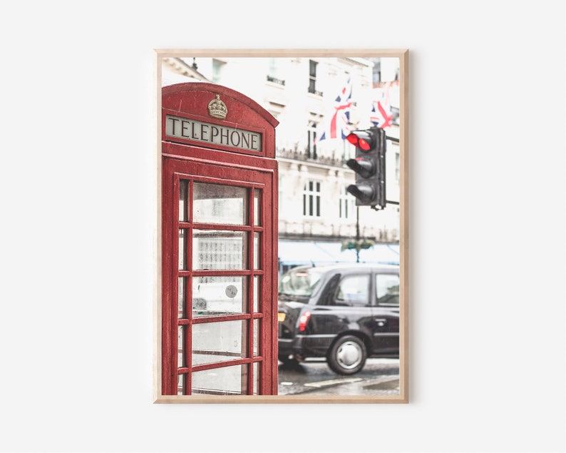 May include: A red telephone booth with the word "TELEPHONE" on the front, located on a street in London, England. A black taxi cab is driving past the booth, and there is a traffic light in the background.