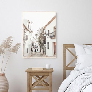 May include: A photograph of a narrow, white-washed street in a European city. The street is lined with buildings that have balconies and windows. The street is empty, and the sun is shining.