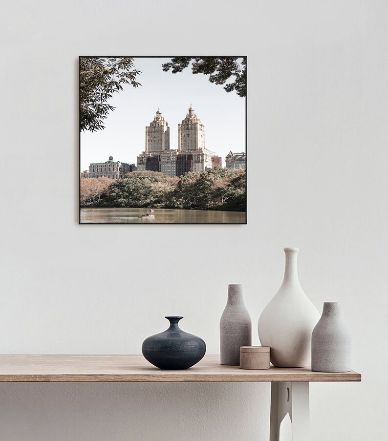 May include: A framed photograph of a city skyline with two tall buildings in the center. The buildings are surrounded by trees and a body of water. The photo is in a black frame and is hanging on a white wall.