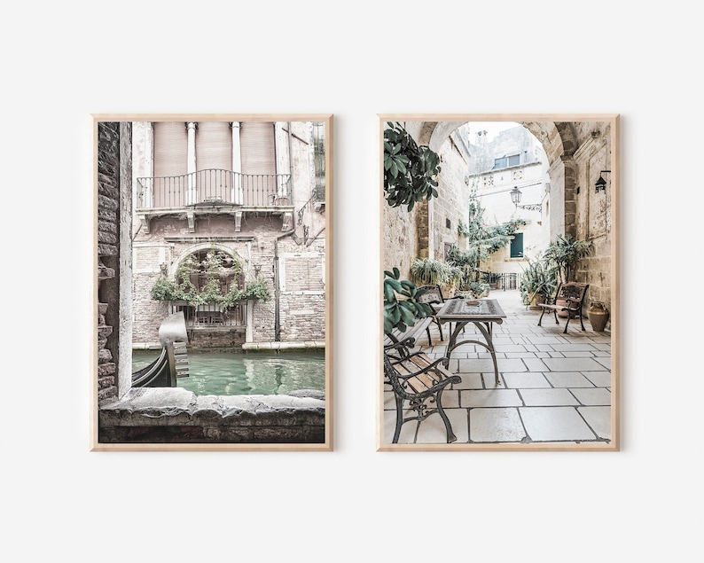 May include: Two framed art prints. The left print shows a Venetian canal scene with a gondola. The right print depicts a courtyard with tables, chairs, and lush greenery. Both prints have a neutral color palette with a vintage aesthetic.