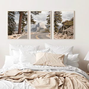 May include: Three framed landscape prints featuring a winding road, towering trees, and a mountain range under a cloudy sky. The prints are displayed above a bed with white linens, a beige pillow, and a knitted throw.