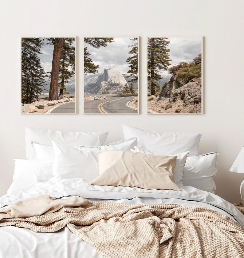 May include: Three framed landscape prints featuring a winding road, towering trees, and a mountain range under a cloudy sky. The prints are displayed above a bed with white linens, a beige pillow, and a knitted throw.