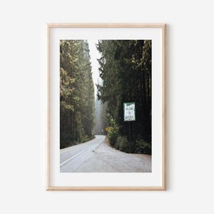 May include: A framed print featuring a scenic road winding through a forest of tall, green trees. A sign that reads "Welcome to Oregon" is visible. The image is framed in a light wood frame.