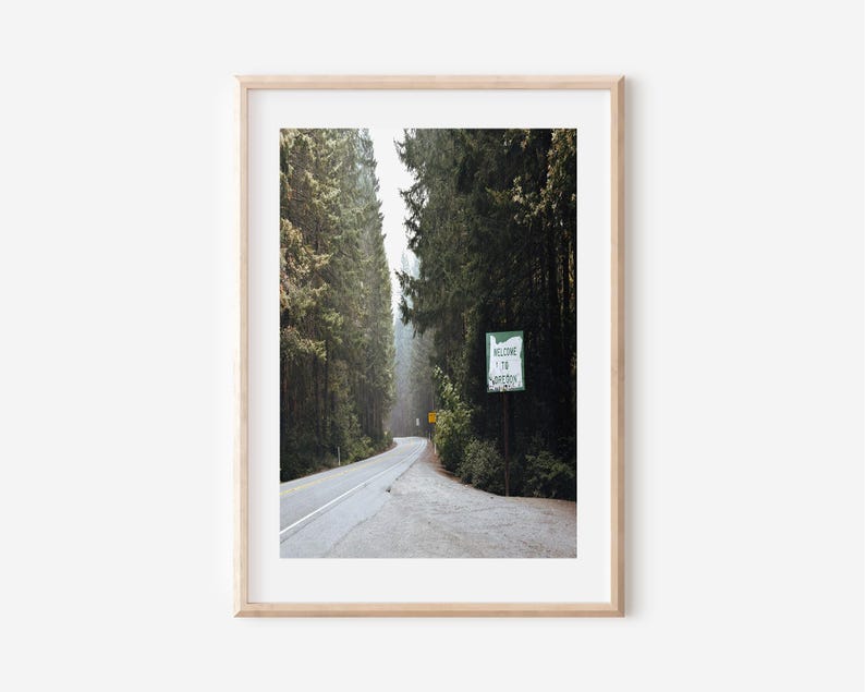 May include: A framed print featuring a scenic road winding through a forest of tall, green trees. A sign that reads "Welcome to Oregon" is visible. The image is framed in a light wood frame.