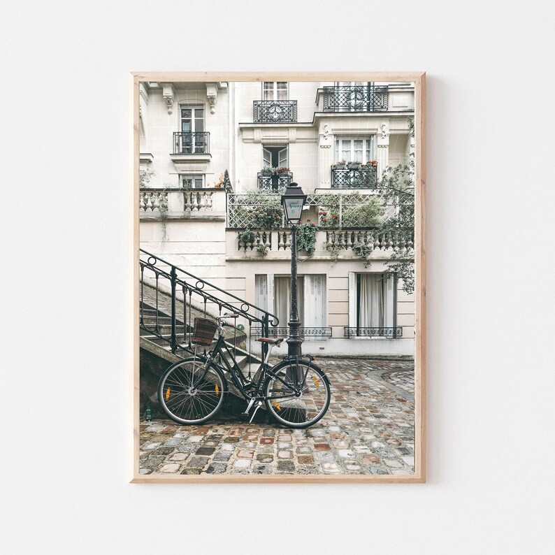 May include: A black bicycle is parked on a cobblestone street in front of a white building with a black wrought iron railing. The building has multiple windows and balconies with plants growing on them. A street lamp is visible in the background.
