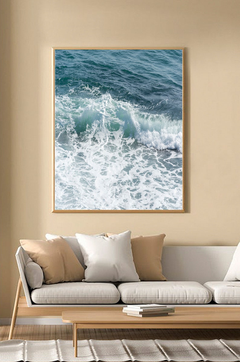 May include: A framed print depicting a breaking ocean wave. The image shows turquoise water forming white foam as it crashes. The artwork is displayed above a white sofa with neutral-coloured cushions.