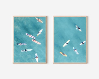May include: Two framed prints of aerial photographs of surfers on turquoise water. The prints are in wooden frames with a light wood finish.
