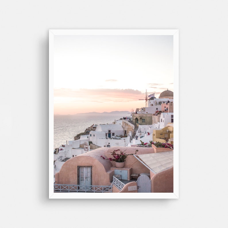 May include: A picturesque view of a whitewashed village in Greece, with a windmill and a domed building in the background. The buildings are clustered together on a hillside overlooking the sea, with a pink sky in the distance.