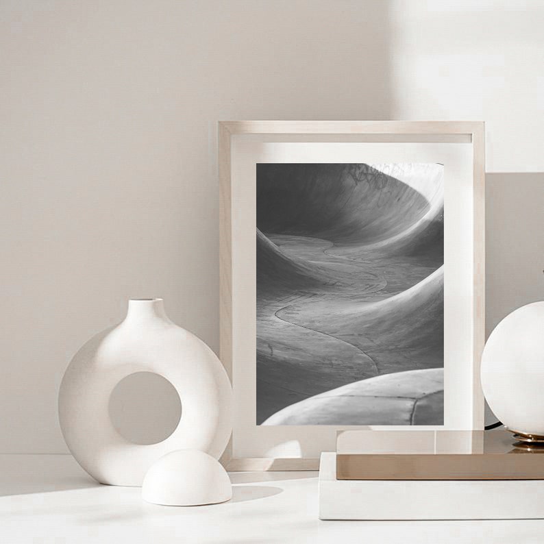 May include: A black and white photograph of a concrete skate park, framed in a white frame. The photo is displayed on a white surface with a white ceramic vase and a white ceramic sphere.