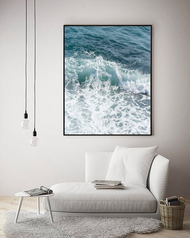May include: A framed print depicting ocean waves in shades of blue and white. The image captures the movement of the water, with a focus on the foamy crests. The artwork is displayed in a modern living space with a white sofa and rug.