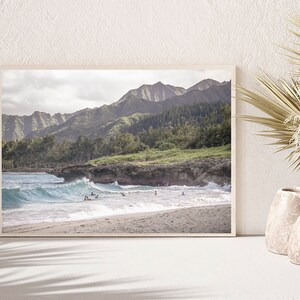 May include: A framed photograph of a beach scene with a mountain range in the background. The beach is sandy with blue water and waves crashing on the shore. There are people in the water, some are surfing.