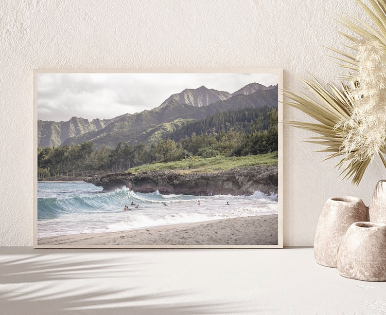 May include: A framed photograph of a beach scene with a mountain range in the background. The beach is sandy with blue water and waves crashing on the shore. There are people in the water, some are surfing.
