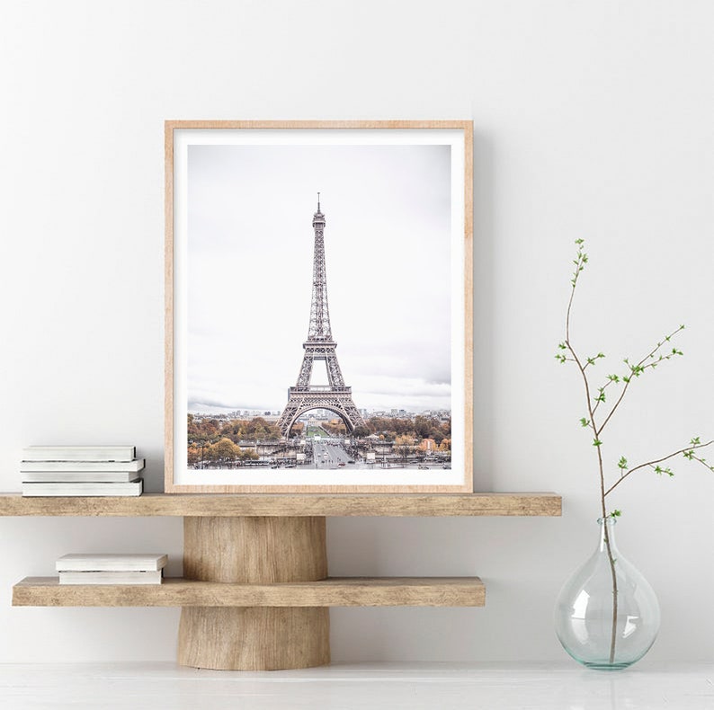 May include: A framed photograph of the Eiffel Tower in Paris, France. The tower is a metal structure with a light gray color. The photo is taken from a distance, showing the tower against a cloudy sky. The photo is framed in a light wood frame.