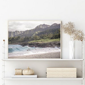 May include: A framed photograph of a beach scene with a mountain range in the background. The beach is sandy with blue water and waves crashing on the shore. There are people in the water surfing.