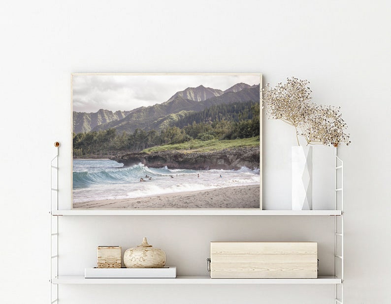 May include: A framed photograph of a beach scene with a mountain range in the background. The beach is sandy with blue water and waves crashing on the shore. There are people in the water surfing.