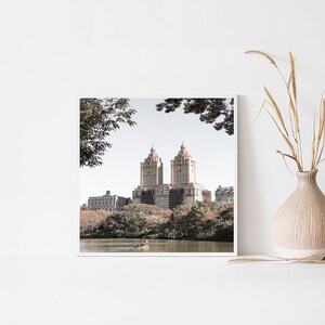 May include: A framed photograph of a cityscape with two tall buildings in the center, surrounded by trees and a lake. The buildings are a light brown color and have a classic architectural style. The trees are a mix of green and brown, and the lake is a calm blue.
