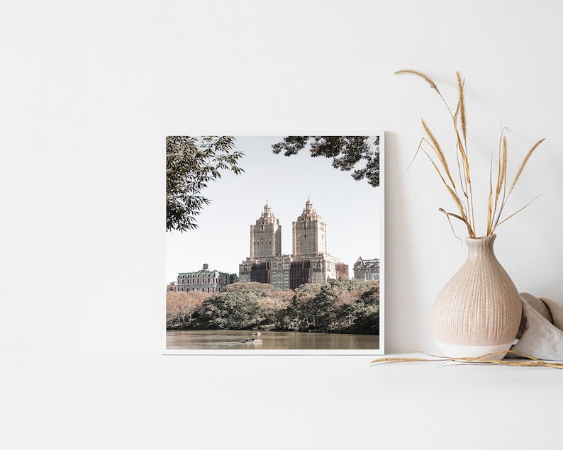 May include: A framed photograph of a cityscape with two tall buildings in the center, surrounded by trees and a lake. The buildings are a light brown color and have a classic architectural style. The trees are a mix of green and brown, and the lake is a calm blue.