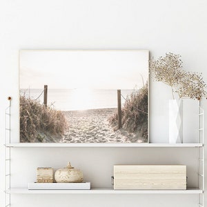 May include: A framed photograph of a beach scene with a wooden walkway leading to the ocean. The photo is in a light brown frame and is hanging on a white wall.