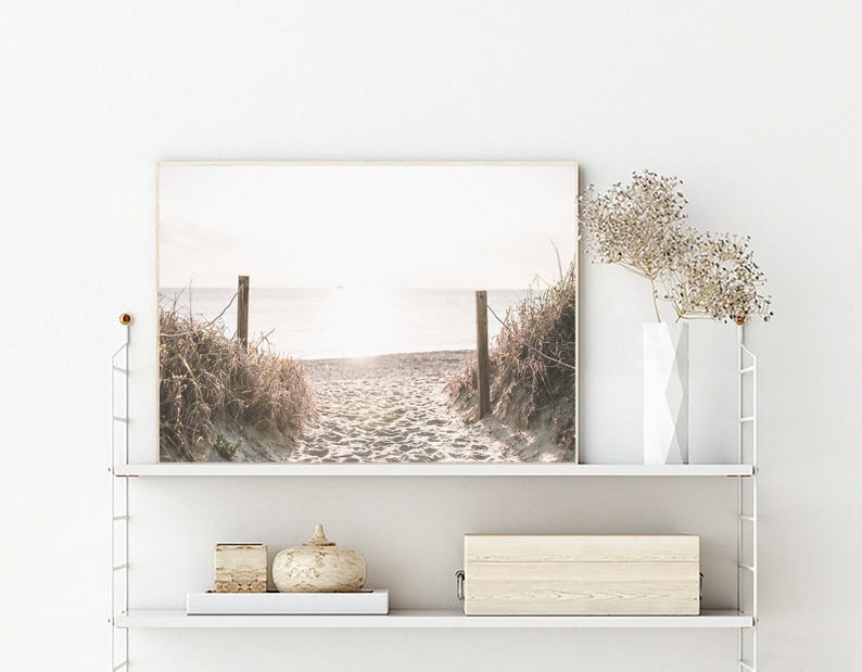 May include: A framed photograph of a beach scene with a wooden walkway leading to the ocean. The photo is in a light brown frame and is hanging on a white wall.