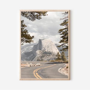 May include: Framed landscape print featuring a winding road leading towards a large, gray mountain. The road has yellow lane markings. The sky is overcast. Trees frame the top of the image, adding a natural border to the scenic view.