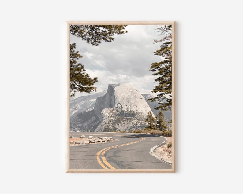 May include: Framed landscape print featuring a winding road leading towards a large, gray mountain. The road has yellow lane markings. The sky is overcast. Trees frame the top of the image, adding a natural border to the scenic view.