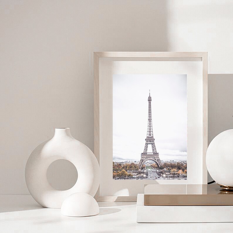 May include: A white ceramic vase with a circular opening in the center, a white ceramic sphere, and a framed photograph of the Eiffel Tower in Paris, France. The photograph is in a white frame with a white mat.