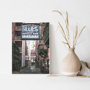 Puede incluir: Un letrero vintage para una banda de blues y un bar boogie de Bourbon Street cuelga sobre un callej&oacute;n estrecho en Nueva Orleans. El letrero es azul con letras blancas y un gr&aacute;fico de teclado de piano. El callej&oacute;n est&aacute; bordeado de edificios de ladrillo y tiene un aspecto desgastado y envejecido.