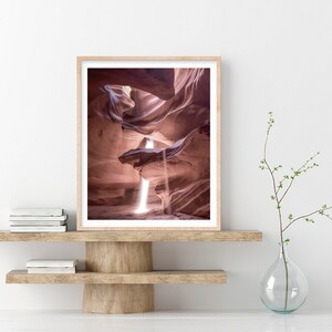 May include: A photograph of a narrow canyon with a beam of light shining through the opening. The canyon walls are a soft pink color and the light creates a dramatic effect.