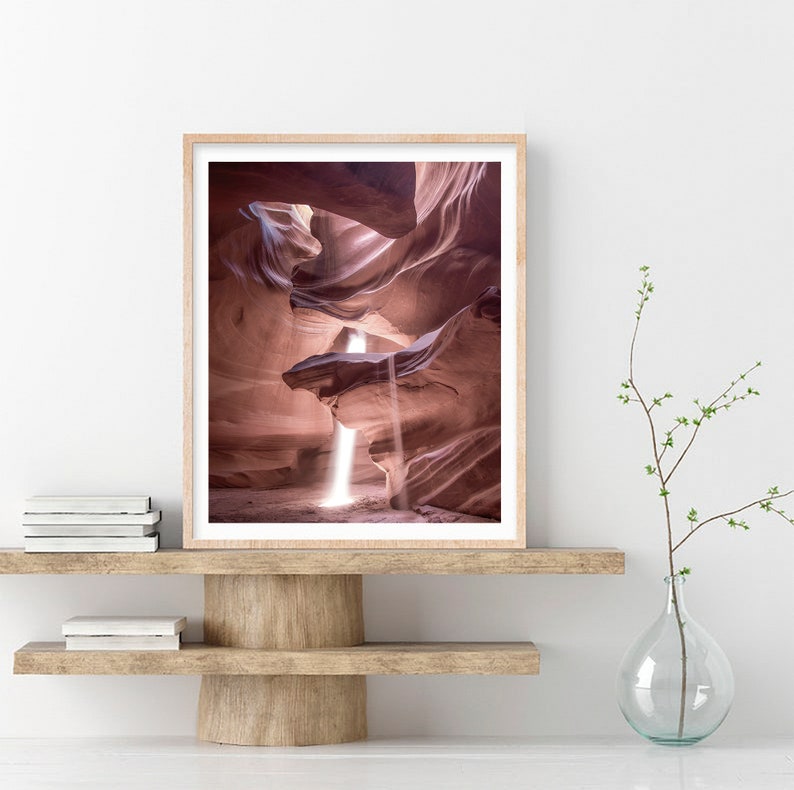 May include: A photograph of a narrow canyon with a beam of light shining through the opening. The canyon walls are a soft pink color and the light creates a dramatic effect.