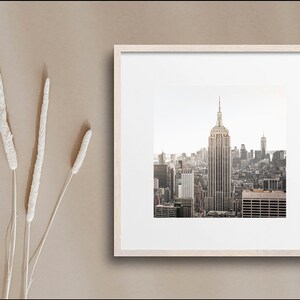 May include: A framed photograph of the New York City skyline with the Empire State Building in the center. The photo is in a light wood frame and is set against a beige background.