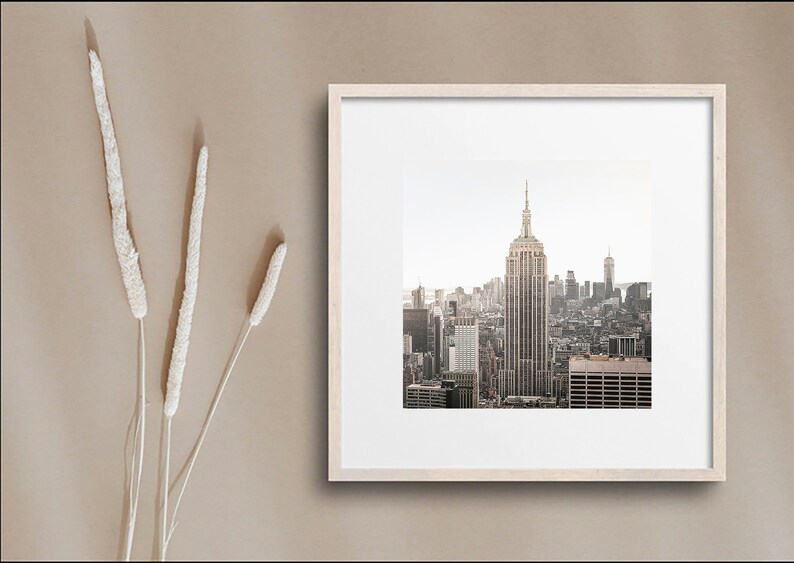May include: A framed photograph of the New York City skyline with the Empire State Building in the center. The photo is in a light wood frame and is set against a beige background.