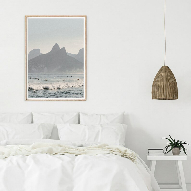 May include: A framed photograph of a beach scene with surfers in the water and mountains in the background. The water is a light blue color and the mountains are a dark gray color. The sky is a light blue color.