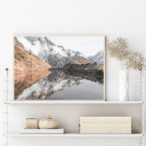 Puede incluir: Una fotograf&iacute;a enmarcada de un lago de monta&ntilde;a con picos cubiertos de nieve que se reflejan en el agua. El lago est&aacute; rodeado de colinas verdes y &aacute;rboles. La foto est&aacute; en un marco de madera claro y est&aacute; colgada en una pared blanca.