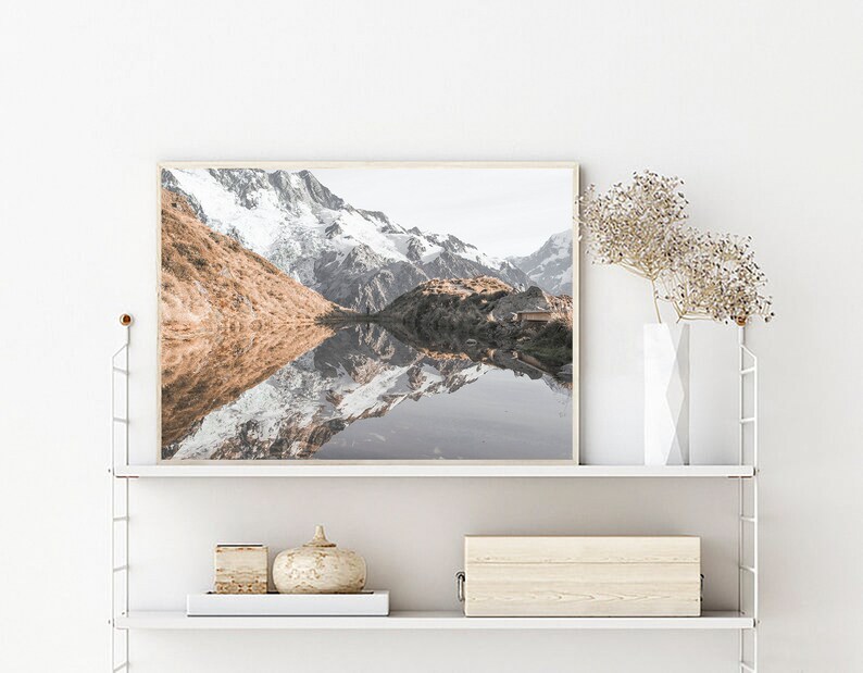 Puede incluir: Una fotograf&iacute;a enmarcada de un lago de monta&ntilde;a con picos cubiertos de nieve que se reflejan en el agua. El lago est&aacute; rodeado de colinas verdes y &aacute;rboles. La foto est&aacute; en un marco de madera claro y est&aacute; colgada en una pared blanca.