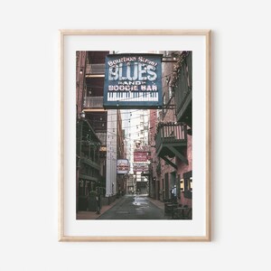 Puede incluir: Un letrero vintage para "Bourbon Street Blues Band and Boogie Bar" cuelga sobre un callej&oacute;n estrecho en un edificio de ladrillo. El letrero es azul con letras blancas y un gr&aacute;fico de teclado de piano. El callej&oacute;n est&aacute; bordeado de edificios de ladrillo y tiene luces de cadena colgando por encima.