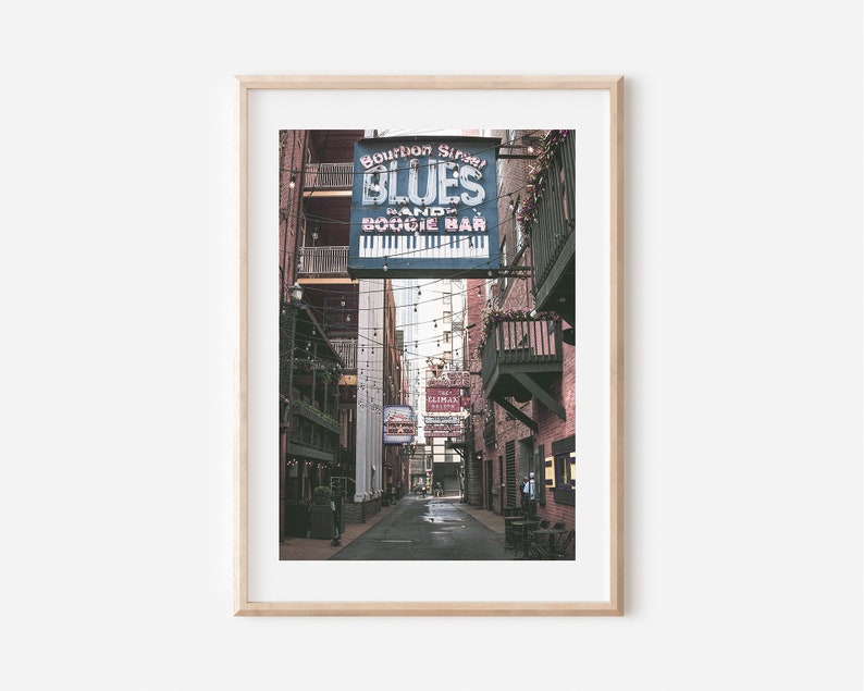 Puede incluir: Un letrero vintage para "Bourbon Street Blues Band and Boogie Bar" cuelga sobre un callej&oacute;n estrecho en un edificio de ladrillo. El letrero es azul con letras blancas y un gr&aacute;fico de teclado de piano. El callej&oacute;n est&aacute; bordeado de edificios de ladrillo y tiene luces de cadena colgando por encima.